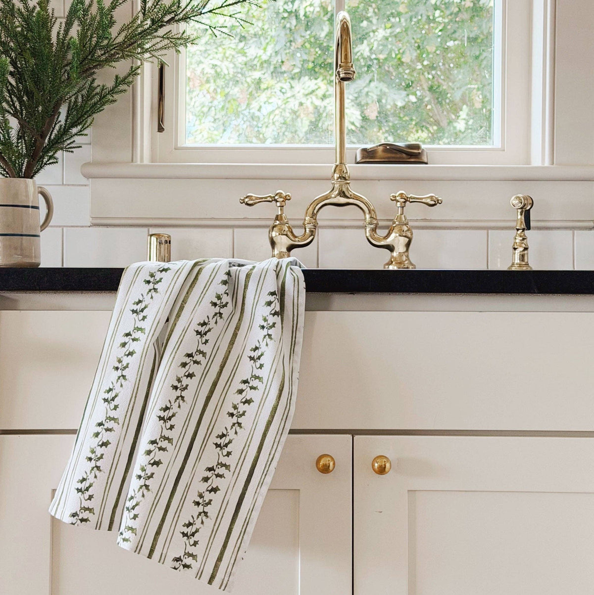 A striped kitchen towel with a green leafy vine pattern hangs from a polished brass farmhouse faucet in a bright, white-tiled kitchen.