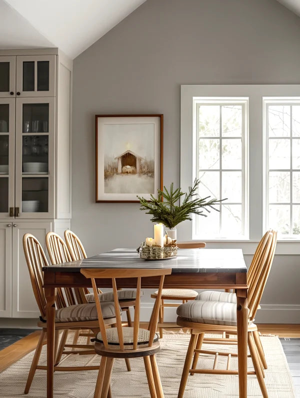 A styled dining room corner featuring a dark wood rectangular table and spindle-back chairs, set on a textured rug against a gray wall with a framed Savior Was Born print and windows.
